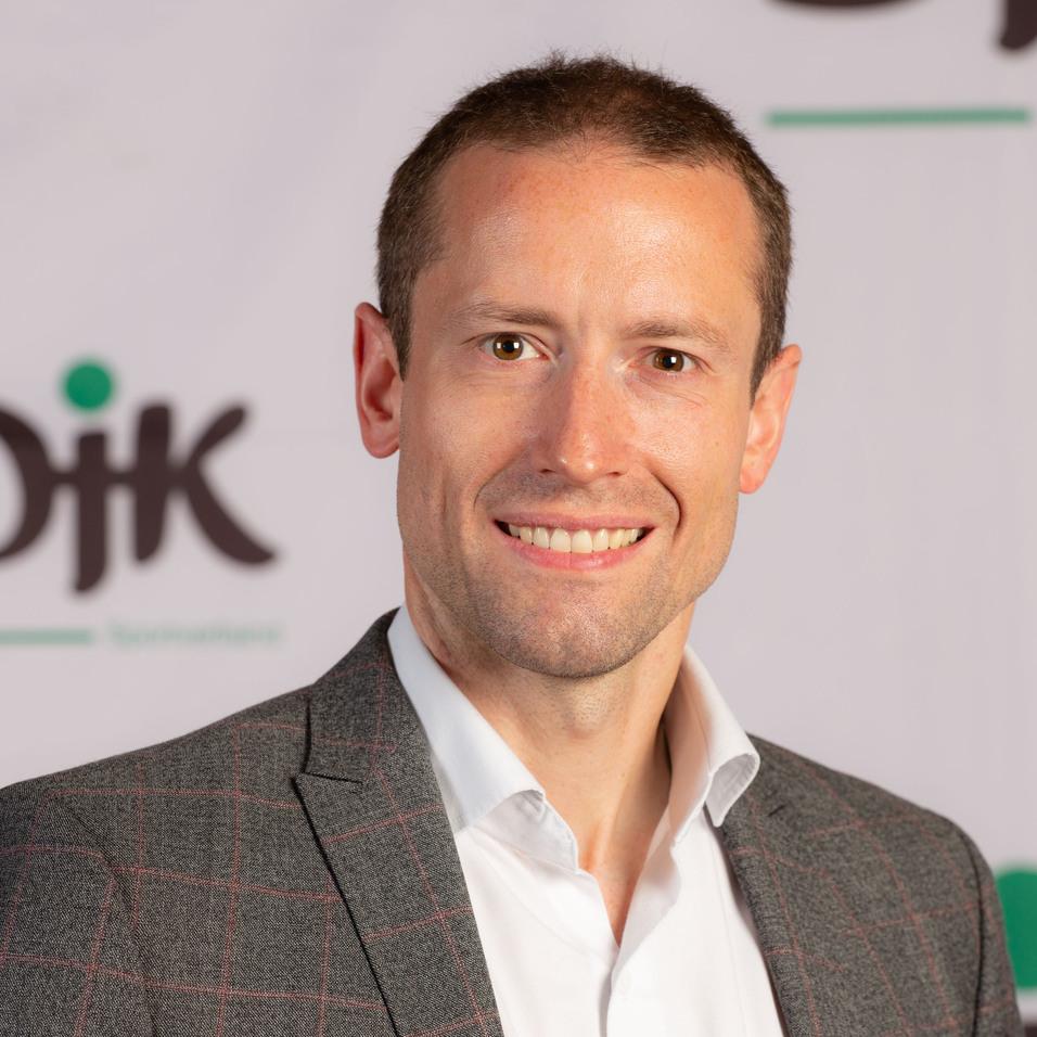 Portr&auml;t eines Mannes in kariertem Blazer vor einem DJK-Logo auf einem hellen Hintergrund. Er l&auml;chelt freundlich.