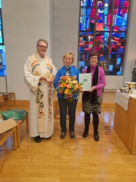 Drei Personen stehen in einer Kirche: ein Geistlicher, eine Frau mit Blumenstrau&szlig; und eine andere Frau mit einer Urkunde.