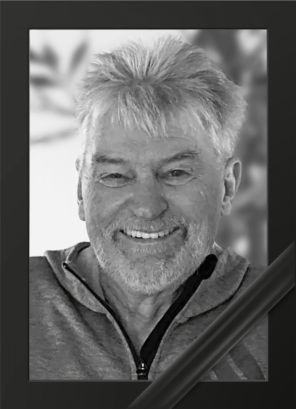 Portr&auml;t eines l&auml;chelnden Mannes mit grauen Haaren in einem grauen Sweatshirt, eingerahmt mit einer schwarzen Trauerschleife.