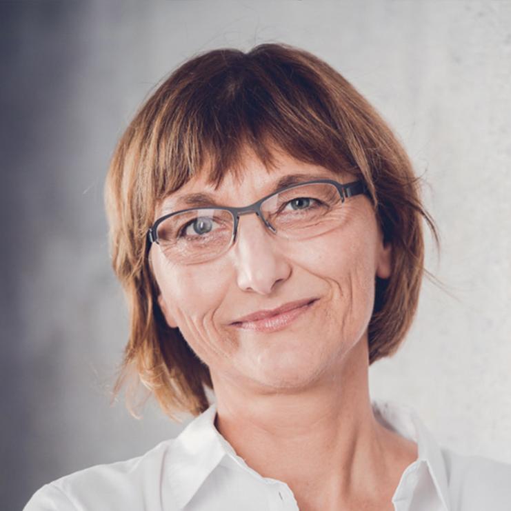 Nahaufnahme einer person mit kurzen braunen Haaren und Brille, die freundlich vor einer hellen Wand l&auml;chelt.