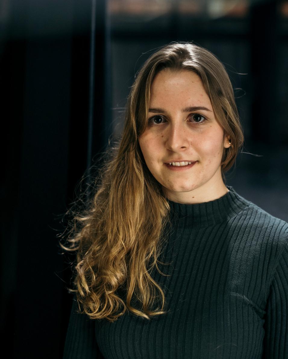 Portr&auml;t einer jungem Frau mit langen, lockigen Haaren, die in einem schlichten, dunkelgr&uuml;nen Pullover l&auml;chelt.