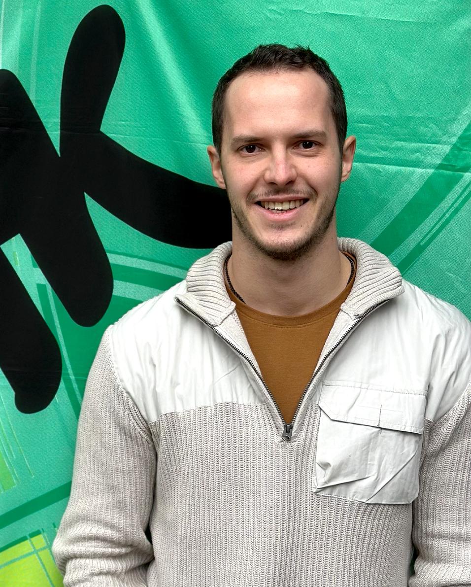 Portr&auml;t eines l&auml;chelnden Mannes in einem hellen Pullover vor einer gr&uuml;nen Hintergrundgrafik.