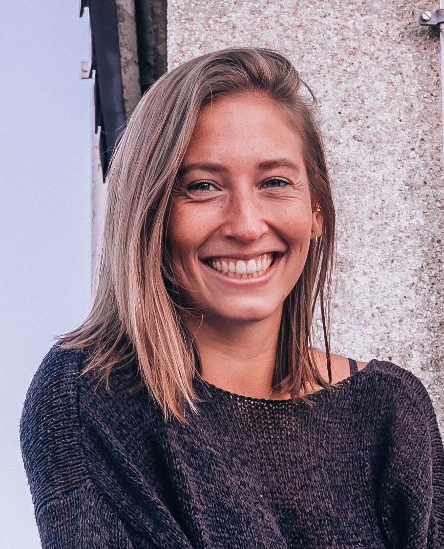 L&auml;chelnde Frau mit mittellangen, blonden Haaren, tr&auml;gt einen dunkelgrauen Pullover, steht vor einer hellen Wand.