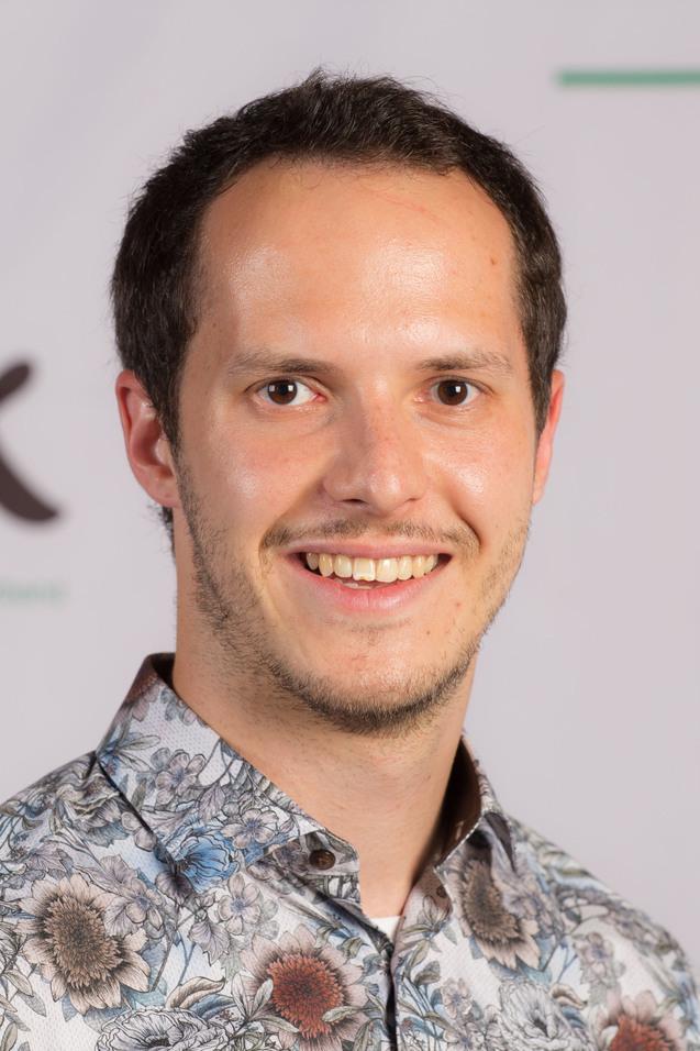 L&auml;chelnder Mann mit kurzen Haaren in einem blumigen Hemd vor einem DJK-Logo auf einer neutralen Hintergrundwand.