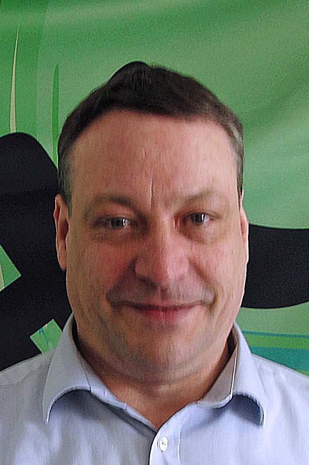 Portr&auml;t eines Mannes mit kurzem Haar, der in ein gr&uuml;nes Hintergrundmuster l&auml;chelt. Er tr&auml;gt ein hellblaues Hemd.