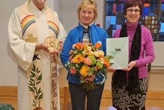 Drei Personen stehen in einer Kirche: ein Geistlicher, eine Frau mit Blumenstrau&szlig; und eine andere Frau mit einer Urkunde.