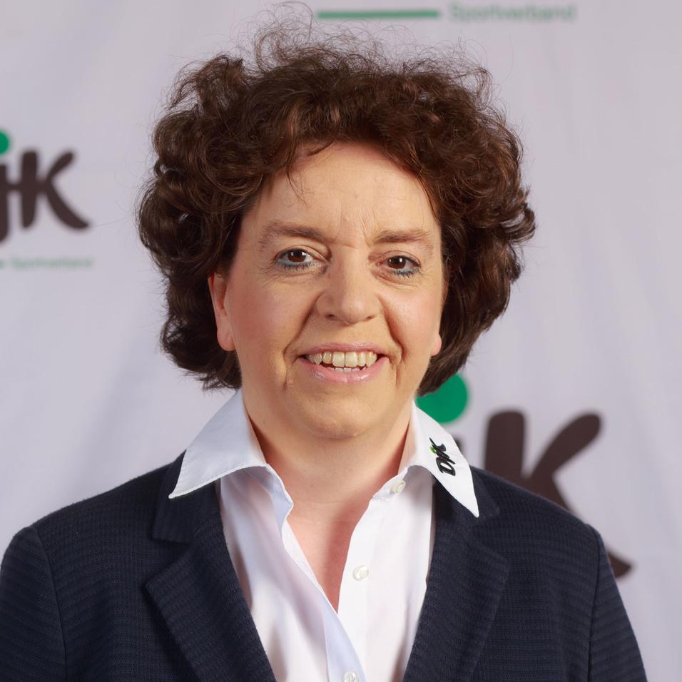 Portr&auml;t einer l&auml;chelnden Frau mit lockigem Haar, gekleidet in einem dunklen Blazer und wei&szlig;em Hemd, vor einem neutralen Hintergrund.