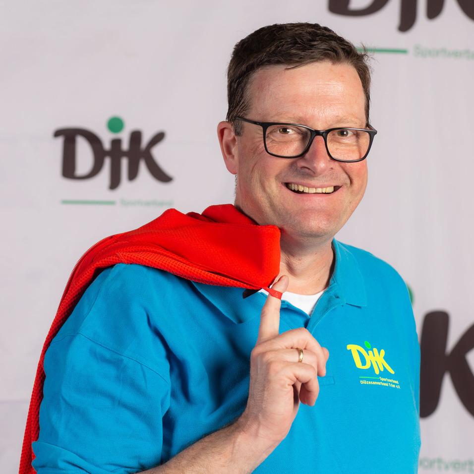 Mann mit Brille und rotem Schal, l&auml;chelt und zeigt mit dem Finger auf sich, vor einem DJK-Hintergrund.