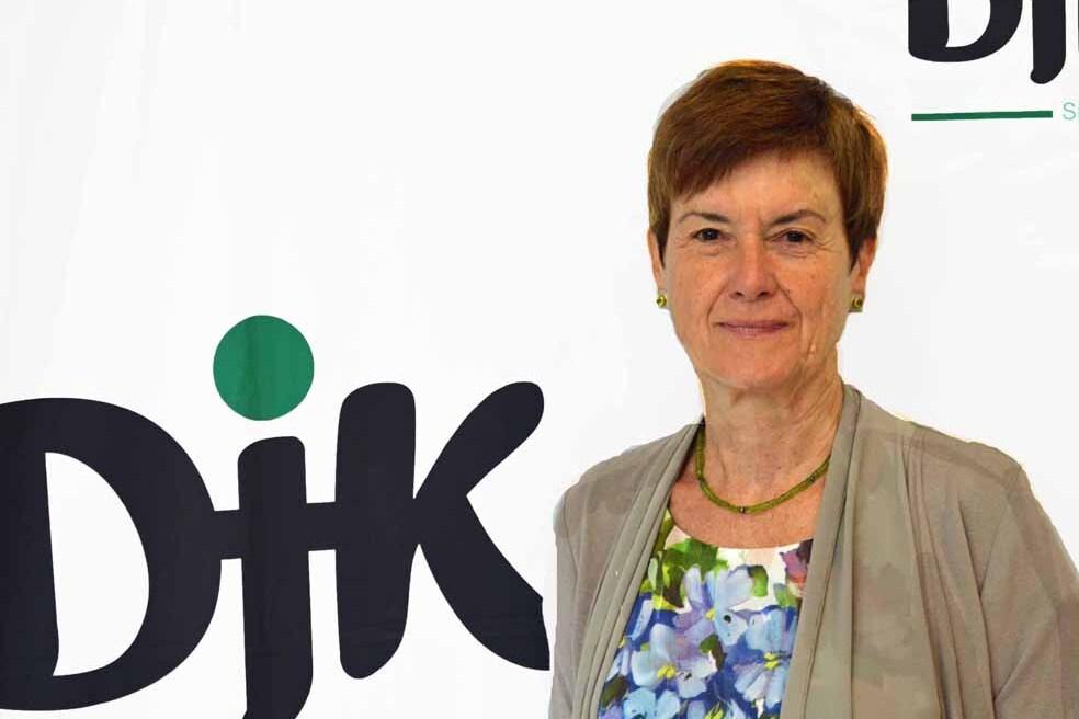 Portr&auml;t einer Frau mit kurzen, braunen Haaren vor einem wei&szlig;en Hintergrund mit dem Logo "DJK" in Schwarz und Gr&uuml;n.