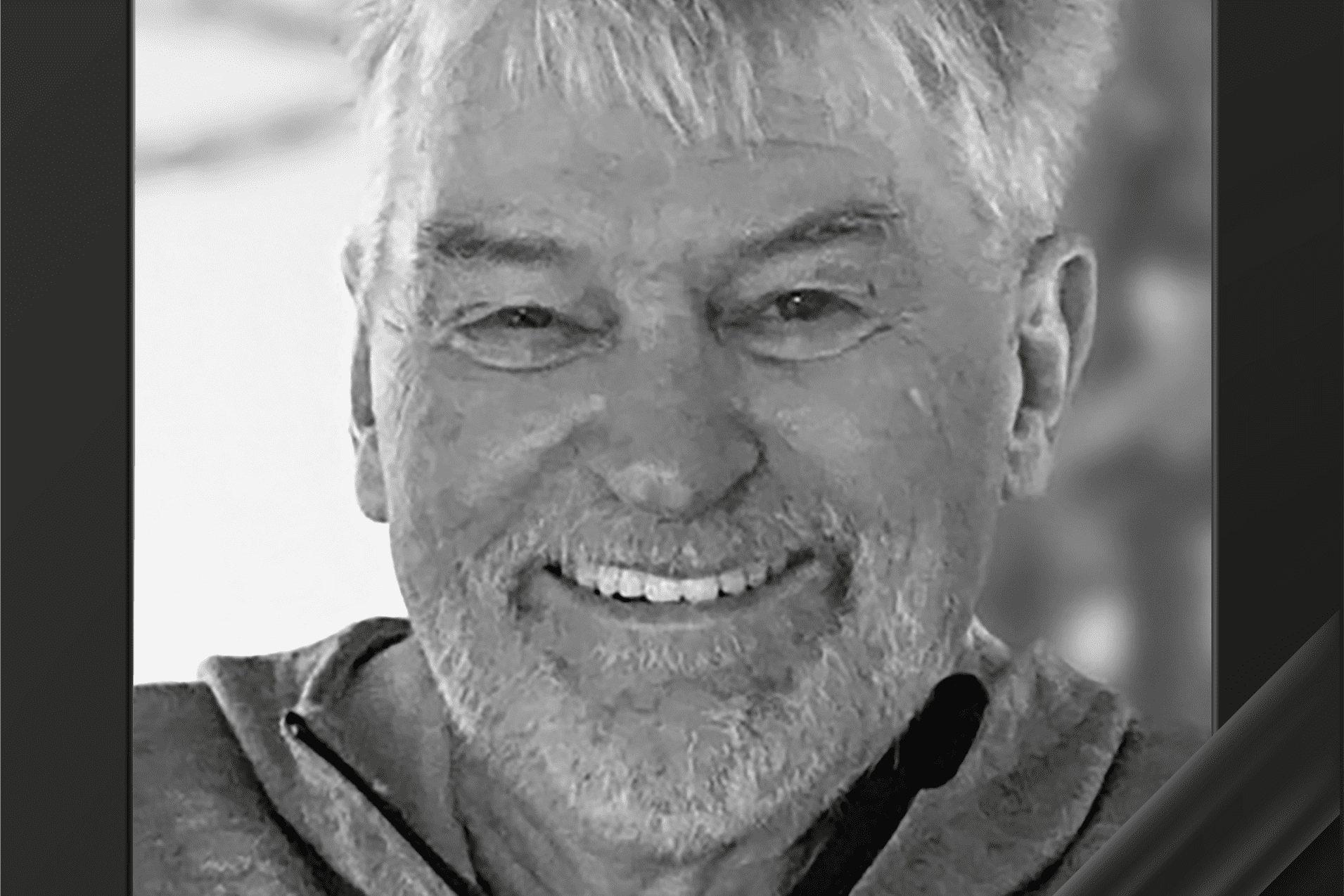 Portr&auml;t eines l&auml;chelnden Mannes mit grauen Haaren in einem grauen Sweatshirt, eingerahmt mit einer schwarzen Trauerschleife.