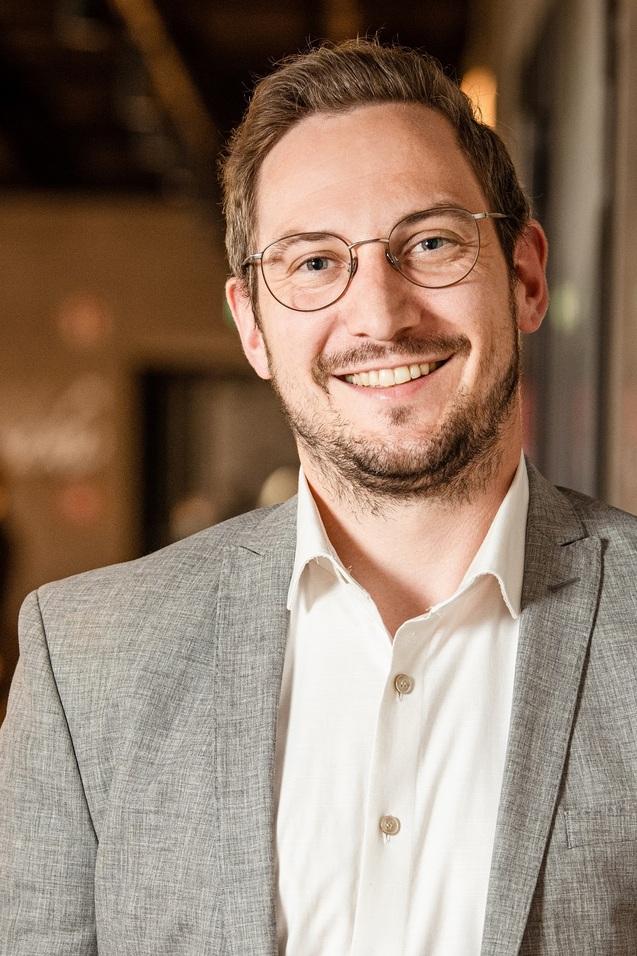 L&auml;chelnder Mann mit Brille und grauem Anzug, steht an einer Wand in einer modernen, hellen Umgebung.