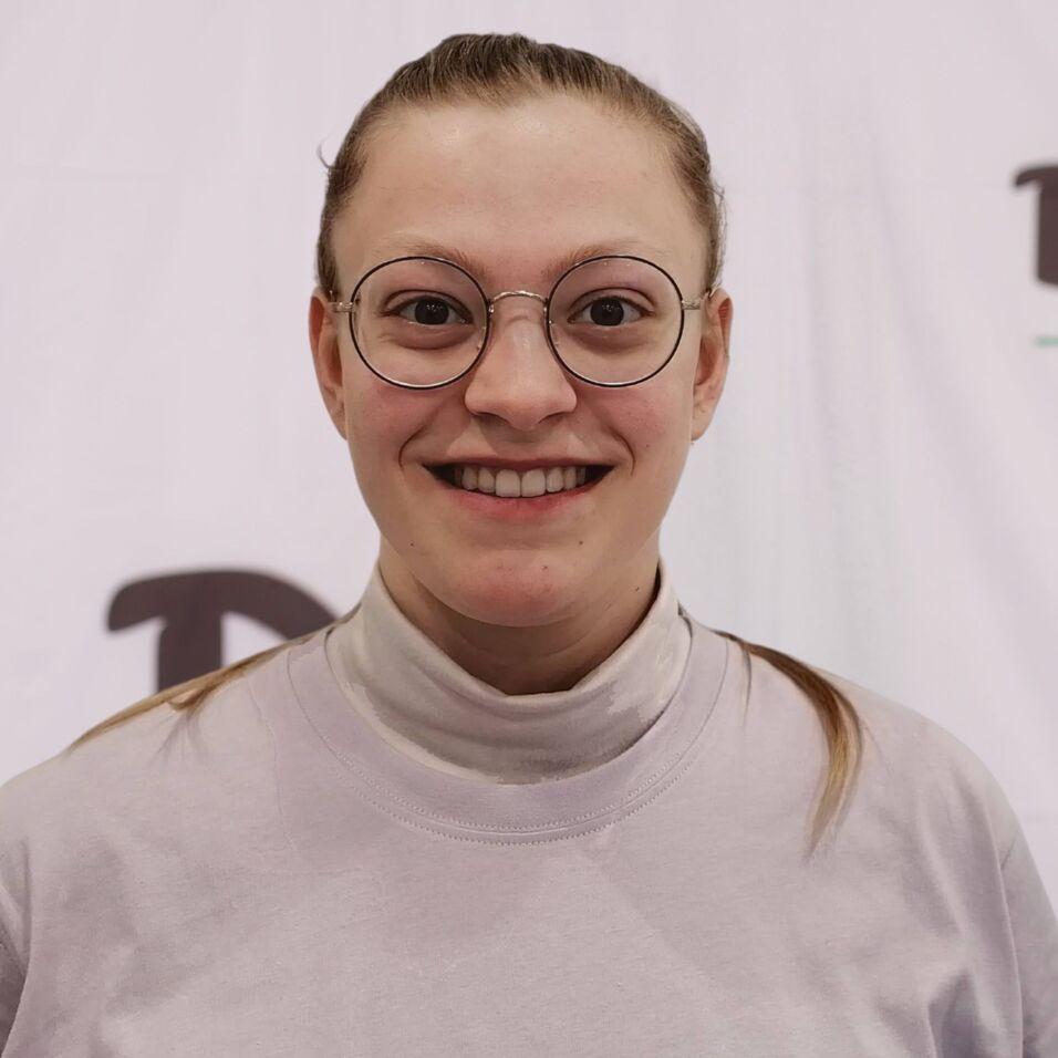 L&auml;chelnde Person mit Brille, tr&auml;gt ein graues Oberteil, vor einem wei&szlig;en Hintergrund mit DJK-Logo.