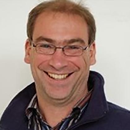 L&auml;chelnder Mann mit Brille und Pullover, vor einem hellen Hintergrund abgebildet.