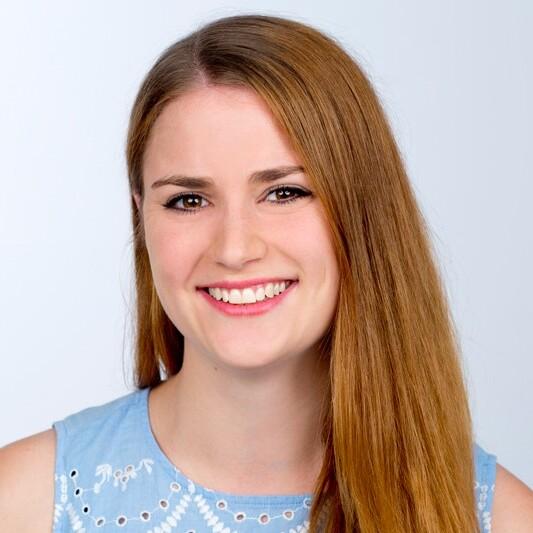 L&auml;chelnde Frau mit langen, glatten Haaren tr&auml;gt ein hellblaues Oberteil mit wei&szlig;en Stickereien vor einem neutralen Hintergrund.