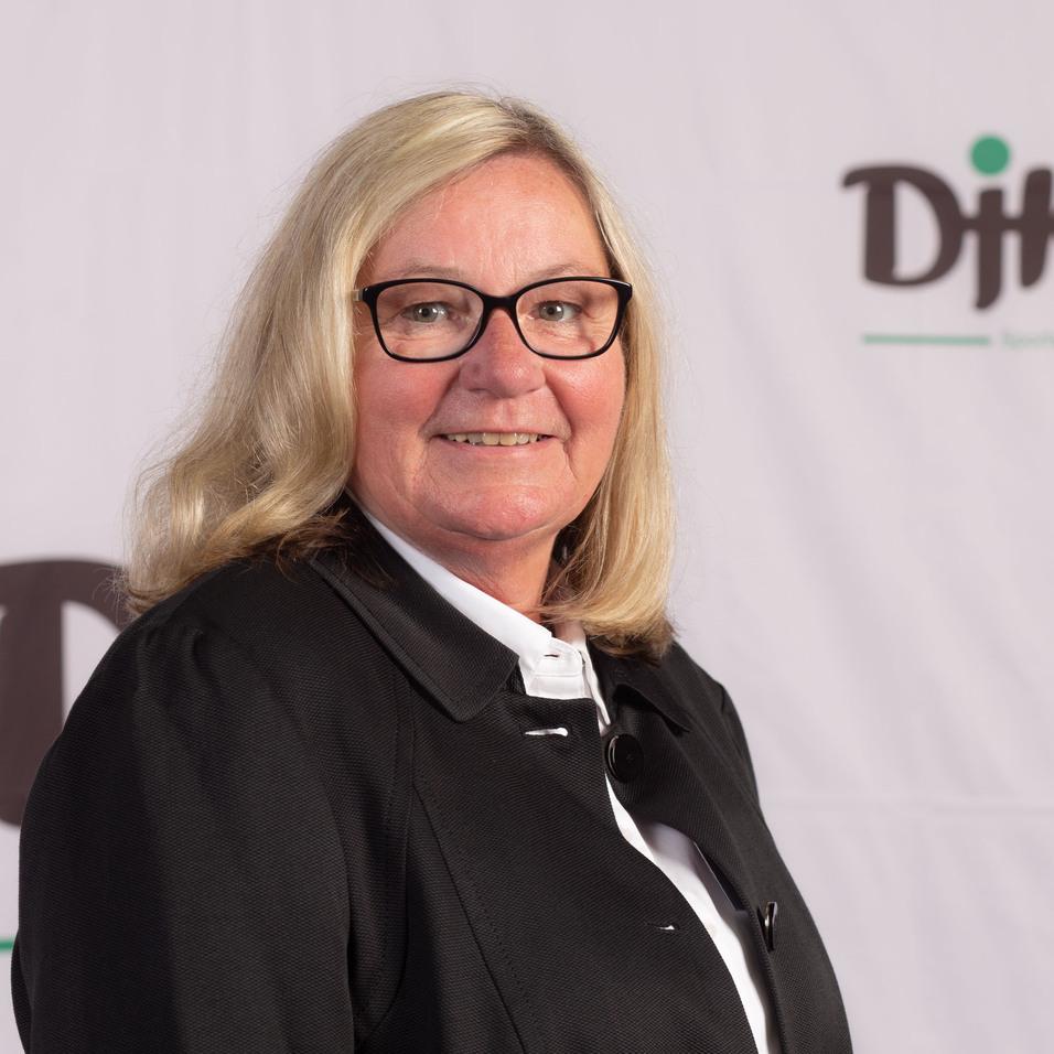 Portrait einer l&auml;chelnden Frau mit blonden Haaren und Brille vor einem DJK-Hintergrund.