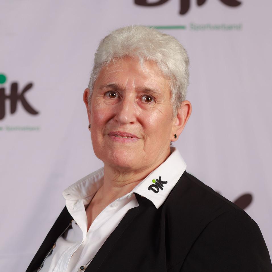 Portr&auml;t einer Frau mit kurzen grauen Haaren, die ein schwarzes Blazer und ein wei&szlig;es Hemd tr&auml;gt, vor einem Logo-Hintergrund.