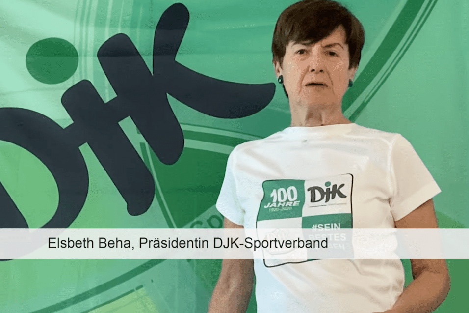 Elsbeth Beha tr&auml;gt ein wei&szlig;es T-Shirt und steht vor einer gr&uuml;nen DJK-Flagge mit dem Text &bdquo;100 Jahre DIK&ldquo;.