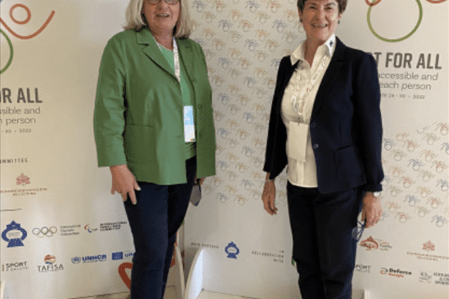 Zwei Frauen stehen l&auml;chelnd vor einer reflektierenden Wand mit dem Slogan &bdquo;Sport for All&ldquo; und sportlichen Symbolen.