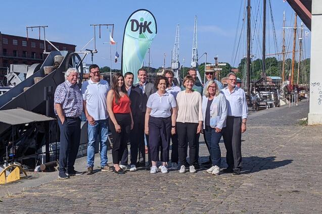 Gruppenfoto von zw&ouml;lf Personen am Hafen, Hintergrund mit Segelbooten und einem gr&uuml;nen DIK-Flaggenbanner.