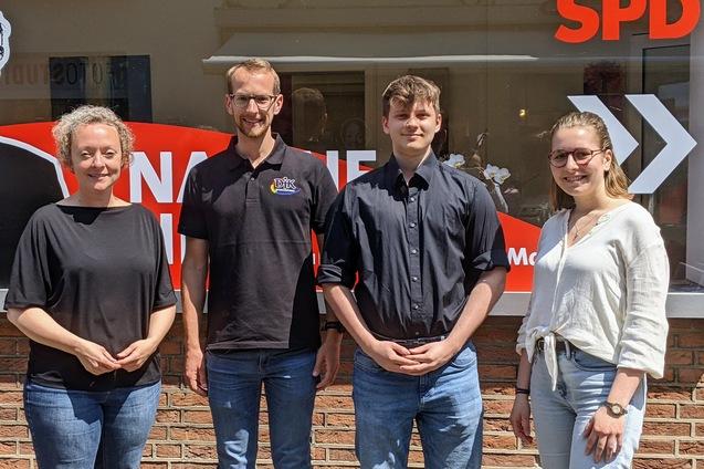 Vier Personen stehen vor einem SPD-B&uuml;ro, l&auml;chelnd, in unterschiedlichen Kleidungsstilen und mit neutralem Hintergrund.