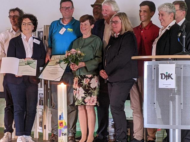 Gruppe von Menschen auf einer B&uuml;hne, feierliche Atmosph&auml;re, eine Person h&auml;lt Blumen und Urkunden.