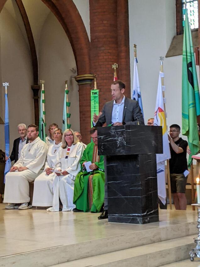 Ein Redner steht an einem schwarzen Rednerpult in einer Kirche, w&auml;hrend versammelte Personen im Hintergrund zuh&ouml;ren.