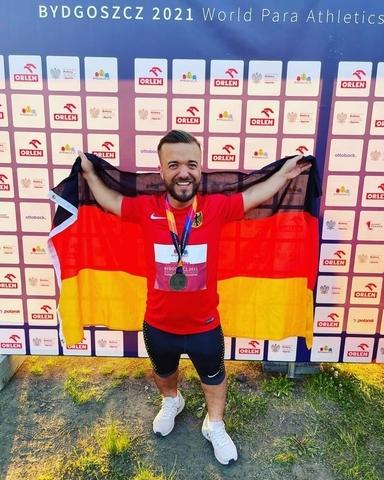 Mann in Sportkleidung h&auml;lt die deutsche Flagge und zeigt seine Medaille vor einer Sponsorenwand.