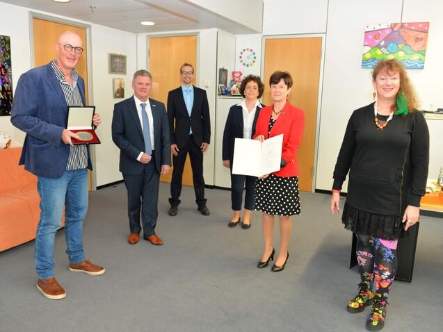 Gruppenszene mit f&uuml;nf Personen in einem B&uuml;ro, zwei halten Urkunden, die anderen beobachten. Das Ambiente ist freundlich und hell.