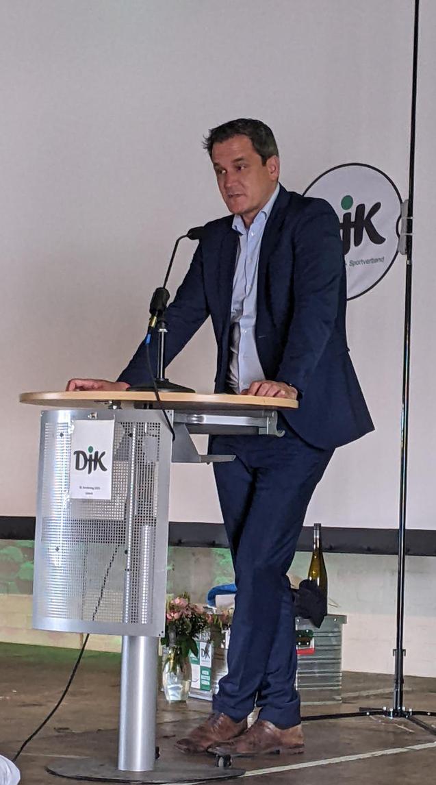 Mann im Anzug steht an einem Rednerpult und h&auml;lt eine Ansprache, im Hintergrund ein Logo und Blumen.