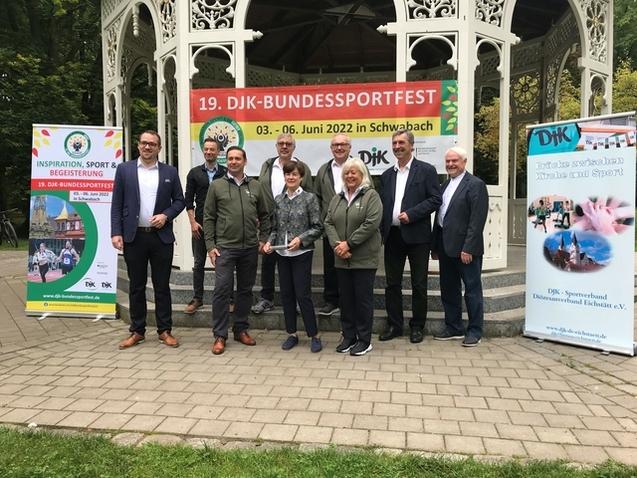 Gruppennahaufnahme von acht Personen vor einem Pavillon, mit Plakaten zum DJK-Bundessportfest im Hintergrund.