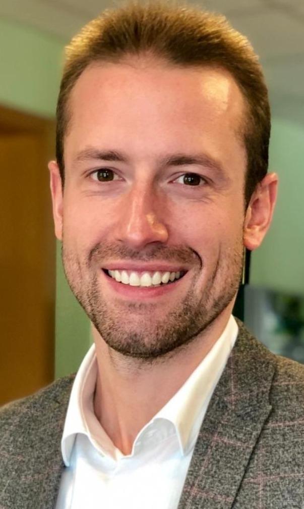 L&auml;chelnder Mann mit kurzen Haaren, grauem Sakko und wei&szlig;em Hemd, vor einem unscharfen Hintergrund in einem B&uuml;ro.