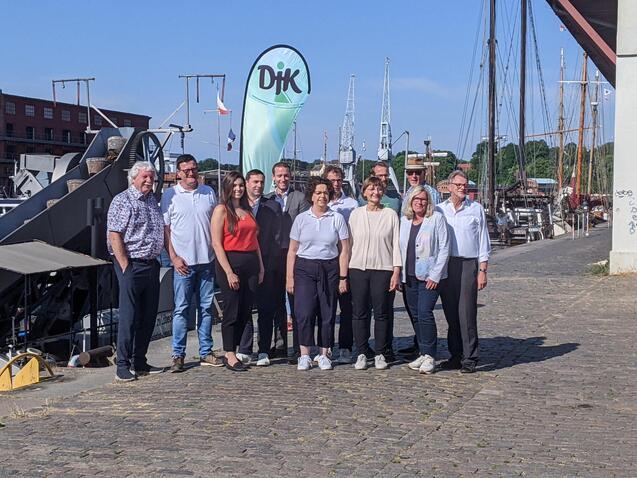 Gruppenfoto von zw&ouml;lf Personen am Hafen, Hintergrund mit Segelbooten und einem gr&uuml;nen DIK-Flaggenbanner.