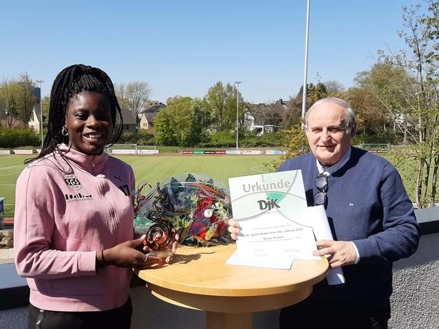 Junge Sportlerin h&auml;lt einen Pokal und eine Urkunde, w&auml;hrend ein &auml;lterer Mann sich mit ihr auf einem Sportplatz fotografieren l&auml;sst.