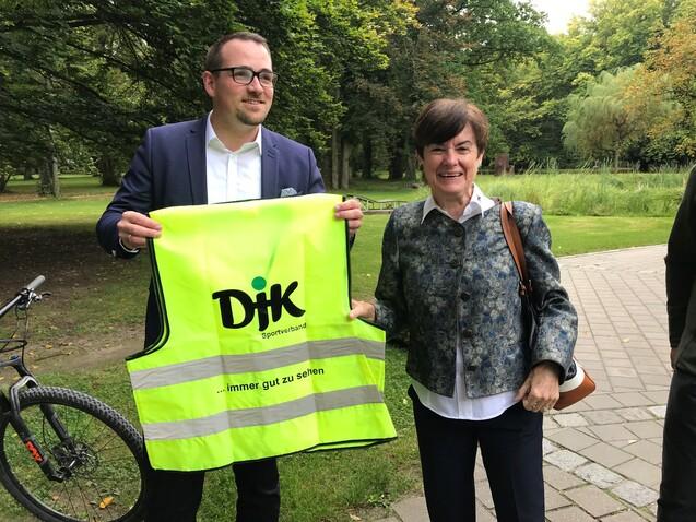Zwei Personen halten eine leuchtend gelbe Warnweste mit dem Aufdruck &bdquo;DJK Sportverband&ldquo; in einem Park.