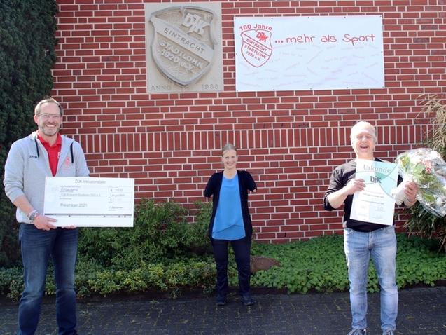 Drei Personen posieren vor einem Geb&auml;ude mit einer Jubil&auml;umsbanner und &Uuml;bergabe von Urkunde sowie Scheck und Blumen.