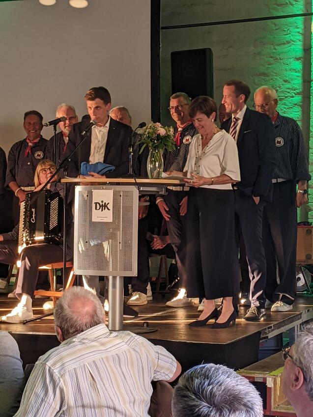 Mann am Mikrofon, w&auml;hrend eine Frau am Pult liest; Menschen im Hintergrund, Blumenstrau&szlig; und Akkordeon sichtbar.