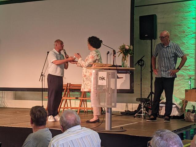 Zwei Personen stehen auf einer B&uuml;hne und unterhalten sich, w&auml;hrend ein dritter Mann im Hintergrund beobachtet.