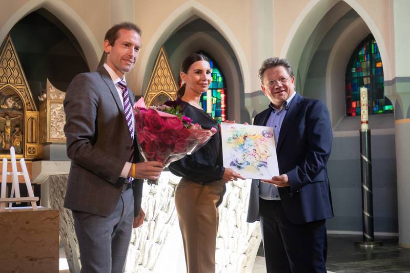 Drei Personen stehen in einer Kirche, eine h&auml;lt einen Blumenstrau&szlig;, eine andere ein Kunstwerk. Buntes Licht strahlt durch die Fenster.