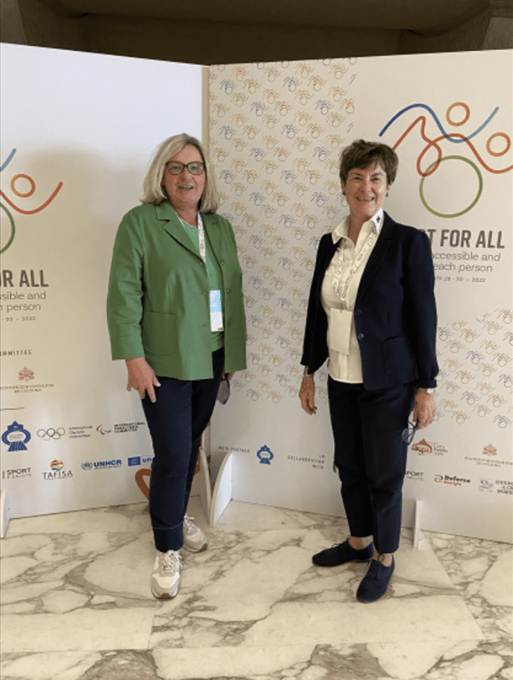 Zwei Frauen stehen l&auml;chelnd vor einer reflektierenden Wand mit dem Slogan &bdquo;Sport for All&ldquo; und sportlichen Symbolen.