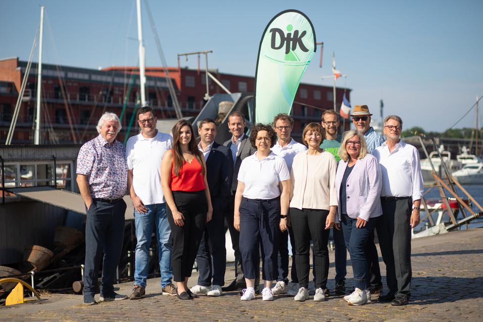 Gruppe von elf Personen am Hafen, Steg mit Booten im Hintergrund, ein gr&uuml;nes Banner mit &bdquo;Djk&ldquo; im Vordergrund.