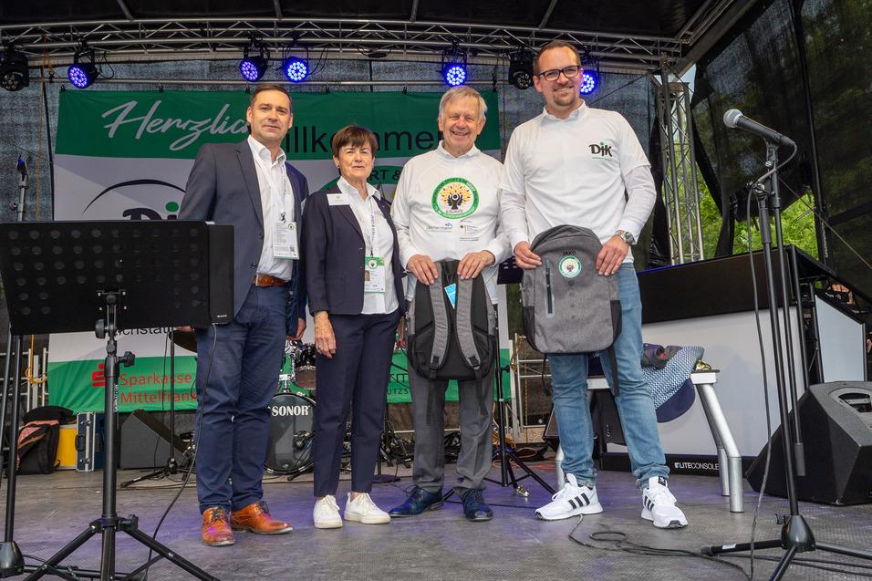 Vier Personen stehen auf einer B&uuml;hne mit Mikrofonen und Banner im Hintergrund, einige halten Rucks&auml;cke in der Hand.