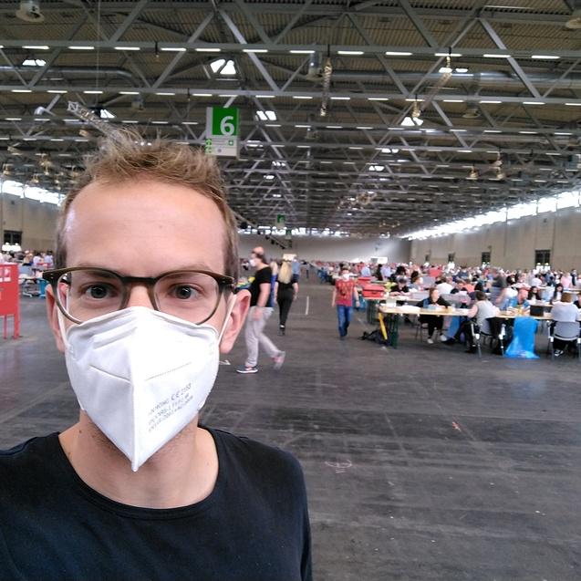 Selbstportr&auml;t einer Person mit Maske, im Hintergrund Menschen an Tischen in einer gro&szlig;en, hellen Halle.