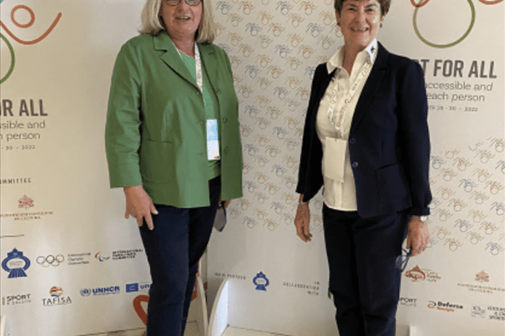 Zwei Frauen stehen l&auml;chelnd vor einer reflektierenden Wand mit dem Slogan &bdquo;Sport for All&ldquo; und sportlichen Symbolen.