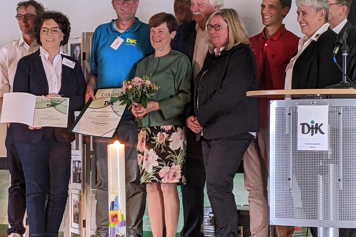 Gruppe von Menschen auf einer B&uuml;hne, feierliche Atmosph&auml;re, eine Person h&auml;lt Blumen und Urkunden.