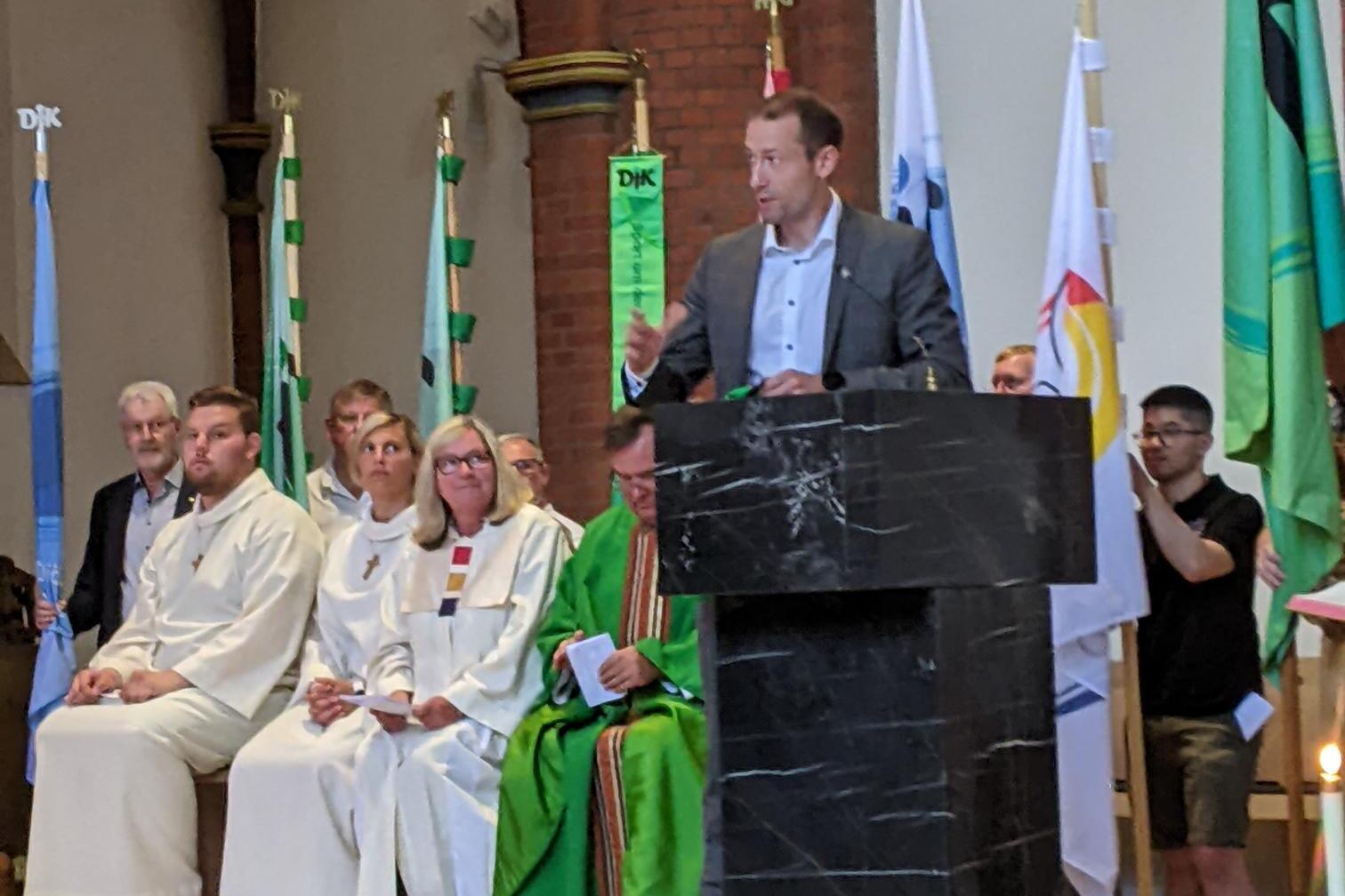 Ein Redner steht an einem schwarzen Rednerpult in einer Kirche, während versammelte Personen im Hintergrund zuhören.