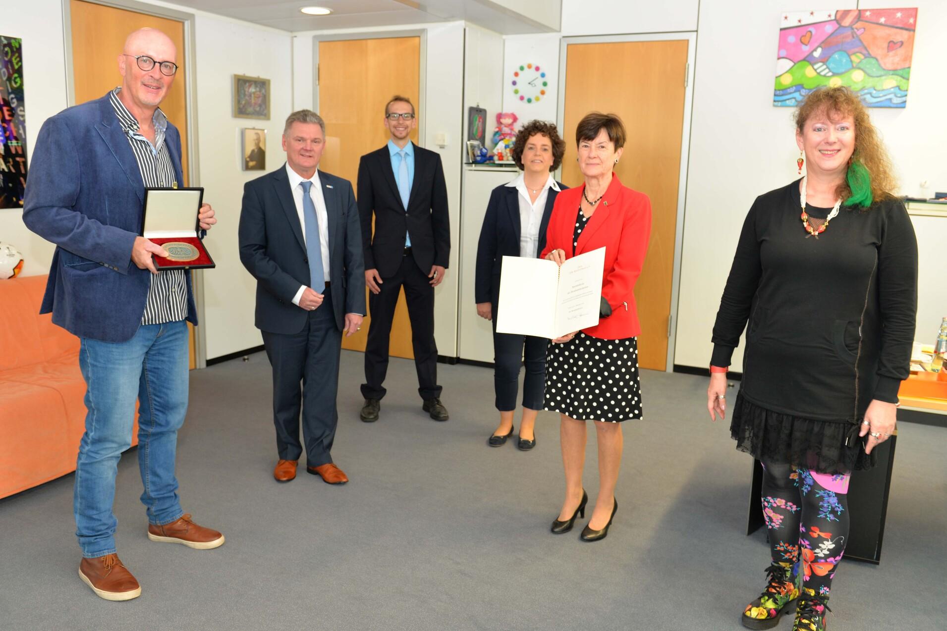 Gruppenszene mit f&uuml;nf Personen in einem B&uuml;ro, zwei halten Urkunden, die anderen beobachten. Das Ambiente ist freundlich und hell.
