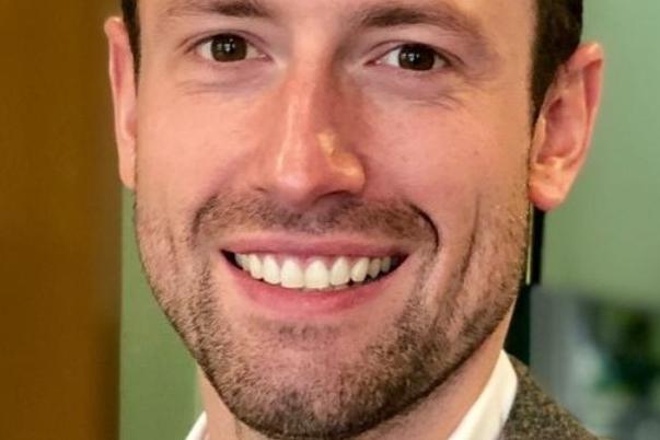 L&auml;chelnder Mann mit kurzen Haaren, grauem Sakko und wei&szlig;em Hemd, vor einem unscharfen Hintergrund in einem B&uuml;ro.