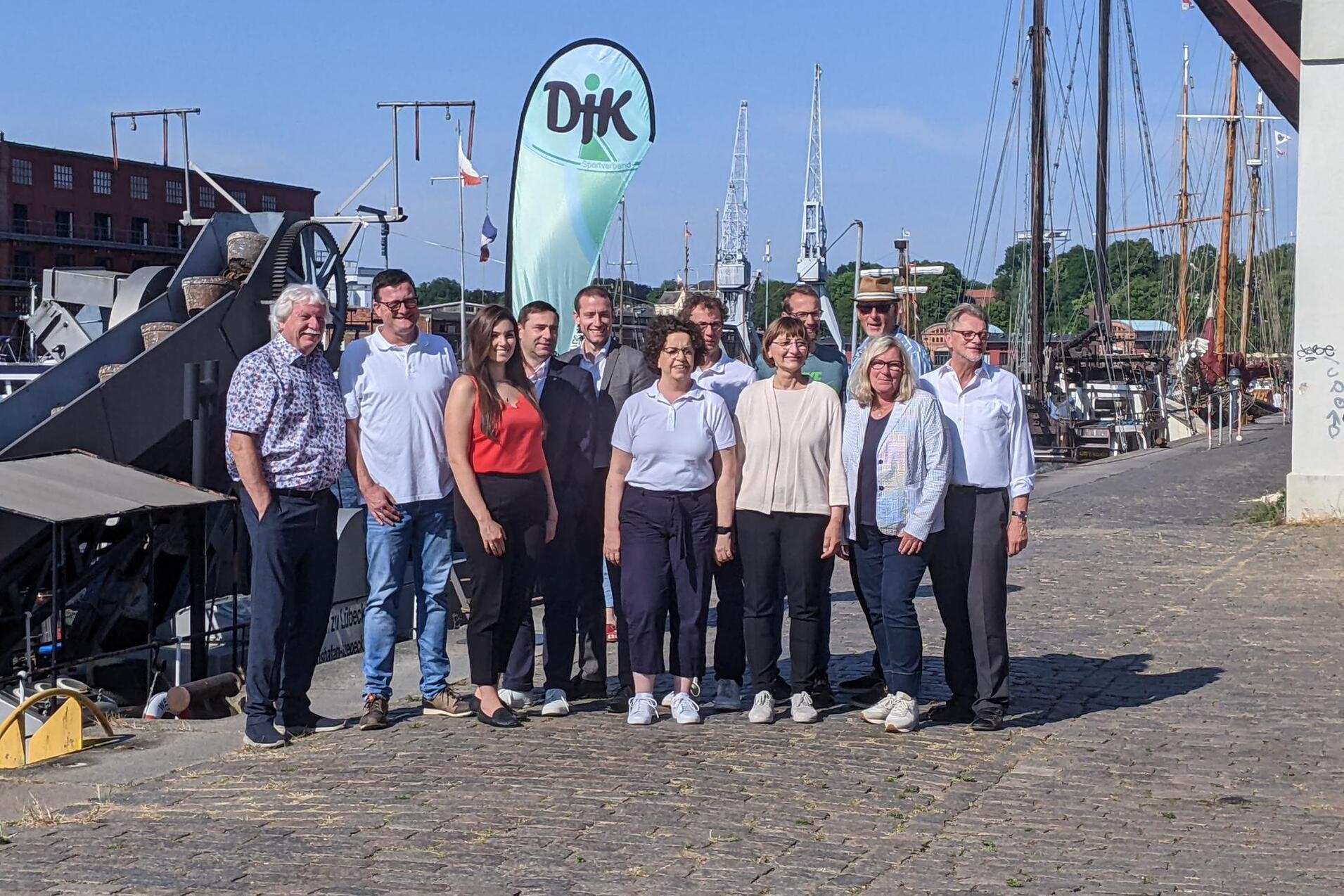 Gruppenfoto von zw&ouml;lf Personen am Hafen, Hintergrund mit Segelbooten und einem gr&uuml;nen DIK-Flaggenbanner.