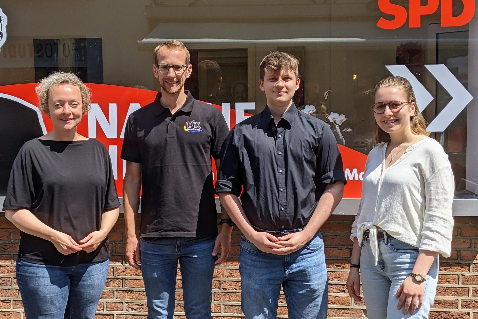 Vier Personen stehen vor einem SPD-B&uuml;ro, l&auml;chelnd, in unterschiedlichen Kleidungsstilen und mit neutralem Hintergrund.