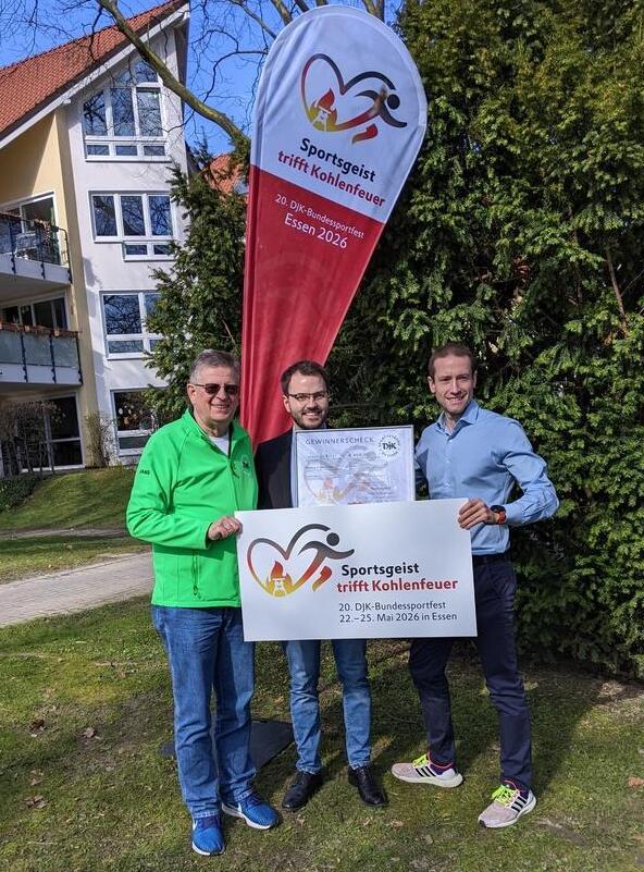 Drei Personen halten ein Banner f&uuml;r das DJK-Bundesportfest 2026, vor einer Baumhecke und einem Geb&auml;ude.
